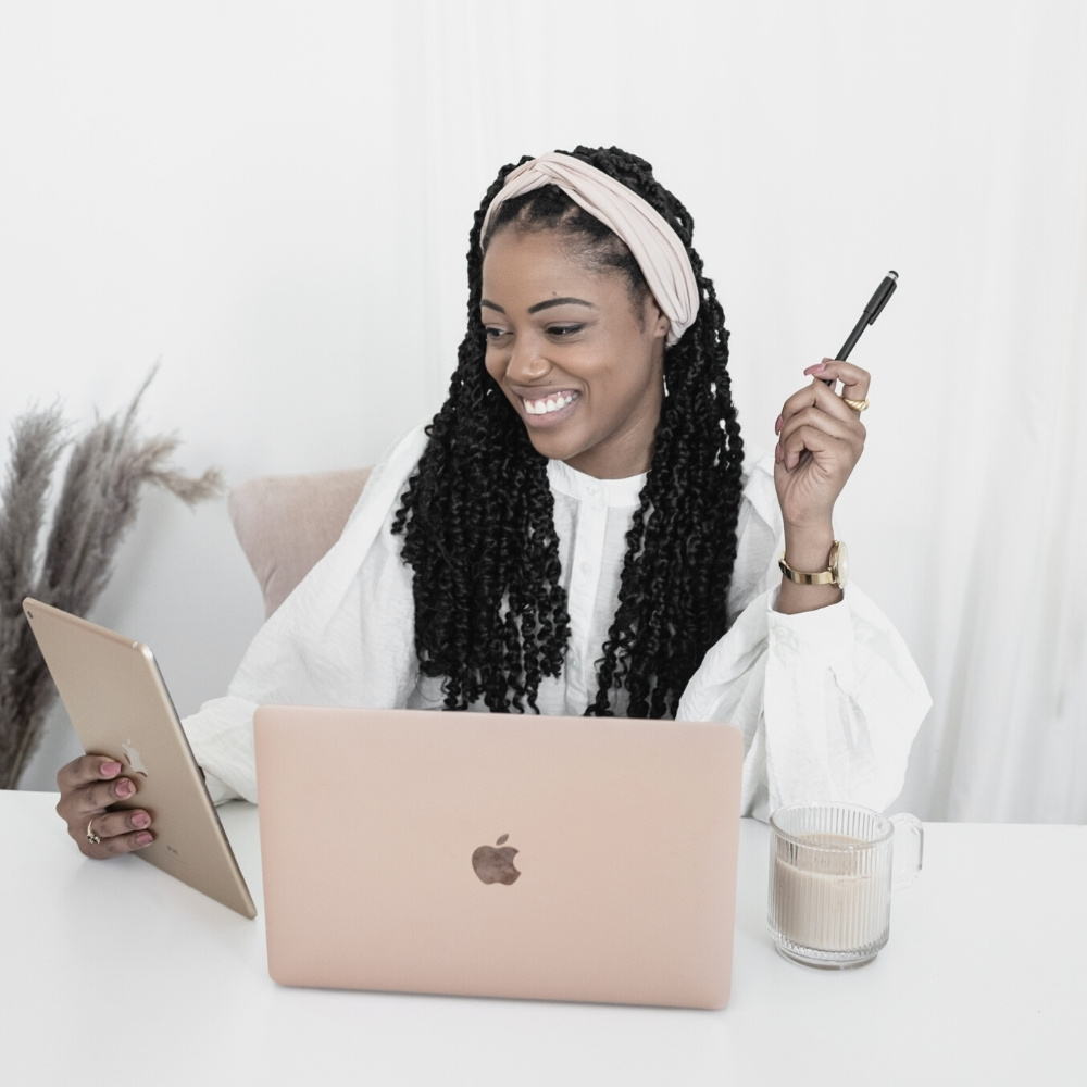 female entrepreneur sitting at her desk with her apple laptop, a cup of coffee while she's holding a pen in her hand and her iPad in the other