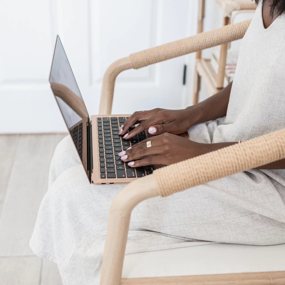 women on a wicker chair typing on rose gold laptop