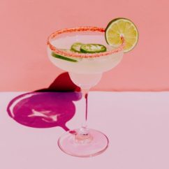 A modern editorial still-life photograph of a spicy margarita positioned on the left side of the composition, shot against a clean neutral background. Strong natural light casts a distinct but soft-edged shadow across the surface, creating visual interest. The drink is a classic pale yellow margarita, with a bold Tajín rim and jalapeño slices clearly visible. The styling is minimal and confident. The image feels graphic, intentional, and fashion-forward, like a magazine opener no people, no text, no borders. --ar 3:4 --profile cdolety --v 7 Job ID: e43d8b86-7960-46fd-bac9-1a92b7250600