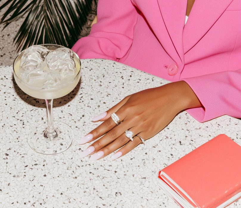 A close-cropped editorial photograph focusing on hands resting on a table, styled with subtle jewelry and polished nails. The surface is off-white with soft texture, while peach-toned elements appear through props like a glass, notebook, or fabric. The subject’s body is only partially visible, creating intrigue and intimacy. Lighting is clean and directional, emphasizing skin texture and material contrast. The composition feels graphic and intentional, like a magazine detail shot that invites the viewer to imagine the rest of the scene. The mood is cool, tactile, and editorial, perfect for a modern brand world. --ar 3:4 --profile 399npew --v 7 Job ID: d39dd78f-80e6-4f6e-bd42-ed4972a6a22b