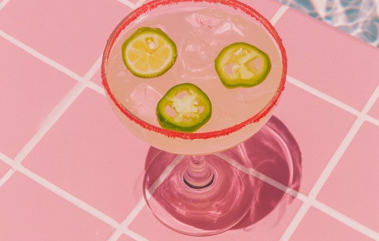 A clean editorial still-life photograph of a spicy margarita resting on pale poolside tile, with soft blue water reflections subtly visible in the background. The margarita is off-center to the left, featuring a deep red-orange Tajín rim, jalapeño slices, and natural condensation. The drink color is classic pale yellow, not green. The composition is minimal and sunlit, with open space to the right. Lighting is bright, high-key daylight, giving a fresh, relaxed mood. The image feels aspirational, calm, and magazine-ready no people, no text, no borders. --ar 3:4 --profile cdolety --v 7 Job ID: 66b45010-66a6-4184-b837-d6d75547e8a9