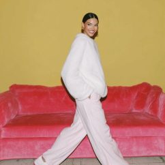 A full-body editorial photograph of a woman walking slowly or standing mid-step in a minimal interior, styled in off-white clothing with a blush accent. The moment feels candid but composed, like an off-duty fashion capture. Her posture is relaxed, with natural movement and a soft, friendly smile. The composition feels effortless and photographic, not staged. Lighting is natural and flattering, enhancing warmth and softness. The image feels cool, approachable, and polished, inspired by Hailey Bieber’s casual editorial moments realistic, full-bleed photography with no borders or frames. --ar 3:4 --profile 399npew --v 7 Job ID: fdfea551-5dc3-4540-9fed-9a544b557891