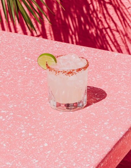 A light editorial still-life photograph of a spicy margarita resting on a simple outdoor table, with the surroundings intentionally minimal and undefined. The margarita is positioned on the left side of the frame, with open negative space. The drink is a traditional pale yellow margarita, with a Tajín chili rim and visible jalapeño slices. Lighting is bright natural daylight, soft and even. The image feels casual, aspirational, and magazine-coded, without feeling staged no people, no text, no borders. --ar 3:4 --profile cdolety --v 7 Job ID: 9a4faf74-2e10-4701-a0e8-d4a146030abc