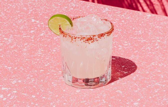 A light editorial still-life photograph of a spicy margarita resting on a simple outdoor table, with the surroundings intentionally minimal and undefined. The margarita is positioned on the left side of the frame, with open negative space. The drink is a traditional pale yellow margarita, with a Tajín chili rim and visible jalapeño slices. Lighting is bright natural daylight, soft and even. The image feels casual, aspirational, and magazine-coded, without feeling staged no people, no text, no borders. --ar 3:4 --profile cdolety --v 7 Job ID: 9a4faf74-2e10-4701-a0e8-d4a146030abc